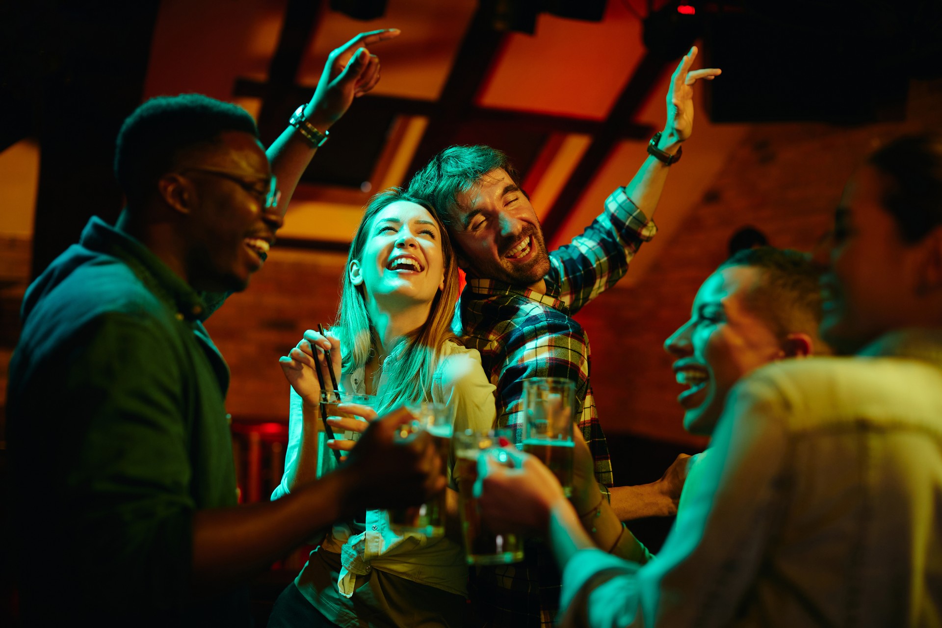 Group of cheerful friends drinking and dancing while gathering in pub at night.